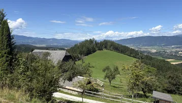 Ländliche Landschaft mit einem Haus, grünen Hügeln und Bergen im Hintergrund unter blauem Himmel.