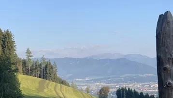 Panoramablick über grüne Hügel und Wälder mit einer Stadt und Bergen in der Ferne unter klarem Himmel.