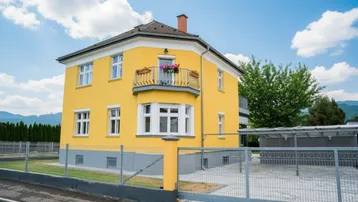 Gelbes Wohnhaus mit weißer Fassade und kleinem Balkon unter blauem Himmel.