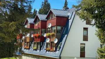 Mehrstöckiges Gebäude mit Satteldach, Gauben und Satellitenschüsseln, umgeben von hohen Bäumen.
