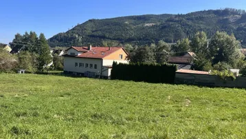 Haus in ländlicher Umgebung mit weitem Grünland und bewaldeten Bergen im Hintergrund unter blauem Himmel.