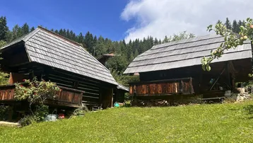 Zwei rustikale Holzhütten mit traditionellen Schindeldächern, umgeben von grünen Wiesen und Bäumen unter blauem Himmel.