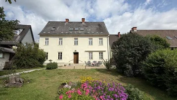 Historisches Gebäude mit gepflegtem Garten, Blumenbeeten und Sitzbereich im Freien unter blauem Himmel.