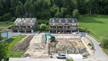 Luftaufnahme einer Baustelle für ein neues Gebäude mit bereits bestehenden Holzhäusern im Hintergrund und grüner Umgebung.
