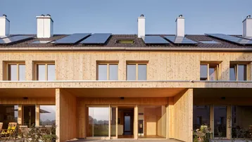 Moderne Holzfassade mit großen Glasfronten, Solarpaneelen auf dem Dach und Zugang zu kleinen Gärten.