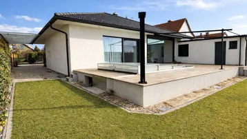 Moderne Hausfassade mit großer Terrasse, Rasenfläche und einer Überdachung, die zum Entspannen einlädt.