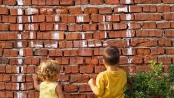 Zwei Kinder zeichnen ein Haus mit Kreide auf eine rote Ziegelwand im Freien.