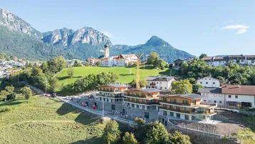 Luftaufnahme eines Neubaukomplexes in einer malerischen Berglandschaft mit traditioneller Kirche im Hintergrund.