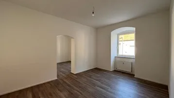 Heller Raum mit Laminatboden und einem Fenster mit Rundbogen, der zu einem weiteren Bereich führt.