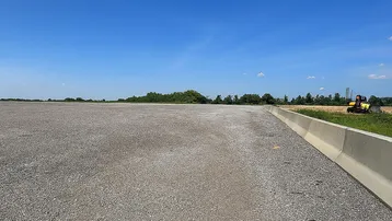 Große, ebene Kiesfläche unter blauem Himmel, umgeben von Grün und mit einem Bagger in der Ferne.