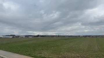 Weitläufige grüne Felder unter einem bewölkten Himmel mit vereinzelten Gebäuden in der Ferne.