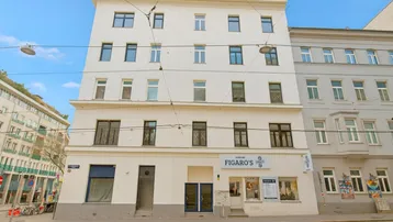 Weißes Mehrfamilienhaus mit klassischer Fassade, vielen Fenstern und einem Geschäft im Erdgeschoss.