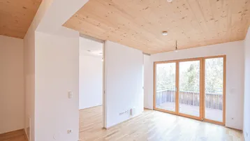 Heller Wohnraum mit Holzboden und Holzdecke, großen Fenstern und Zugang zu einem Balkon.