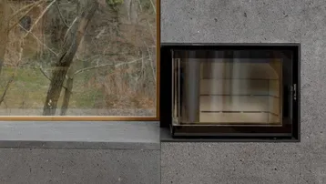 Moderner Kamin aus dunklem Stein mit Holzfußboden und Blick ins Grüne durch das Fenster.