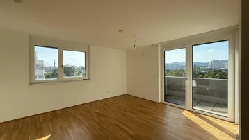 Helles, leeres Wohnzimmer mit Parkettboden und Zugang zu einem Balkon, bietet viel Tageslicht.