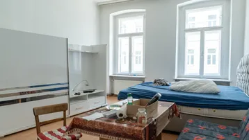 Helles Schlafzimmer mit Bett, Tisch und Schrank, ausgestattet mit zwei großen Fenstern.