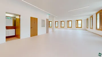 Großer, heller Büroraum mit vielen Fenstern, modernen Leuchten und Zugang zu einem gefliesten Sanitärbereich.