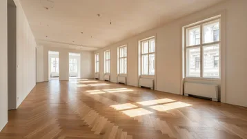 Geräumiger, heller Raum mit Fischgrätparkett und vielen Fenstern, die viel Tageslicht hereinlassen.