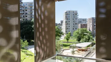 Sonniger Balkon mit perforierten Sichtschutzwänden und weitem Blick auf moderne Gebäude und grüne Parkanlagen.