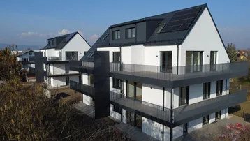 Zwei moderne Mehrfamilienhäuser mit Balkonen und Solaranlagen, eingebettet in eine grüne Umgebung.