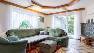 Gemütliches Wohnzimmer mit grünem Sofa, Kachelofen und Zugang zur Terrasse im Freien.