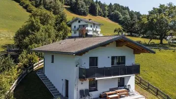 Modernes Haus auf einem Hügel mit Balkon und Terrasse, umgeben von grünen Wiesen und Bäumen.