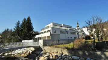 Modernes Gebäude mit Terrassen und Balkonen, gelegen an einem Bach mit Brücke und Blick auf eine Kirche.