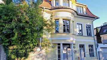 Elegante Stadtvilla mit gelber Fassade, Erker und gepflegtem Vorgarten, umgeben von viel Grün.