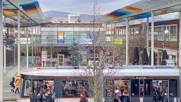 Eine Straßenbahn fährt durch eine moderne Stadtlandschaft mit bunten Überdachungen und Gebäuden im Hintergrund.