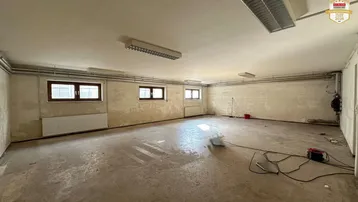 Großer, leerer Raum mit Betonboden, mehreren Fenstern und sichtbaren Heizkörpern. Die Wände zeigen Abnutzungsspuren.
