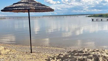 Strandabschnitt mit Strohsonnenschirm und weitem Blick über das ruhige Wasser unter blauem Himmel.