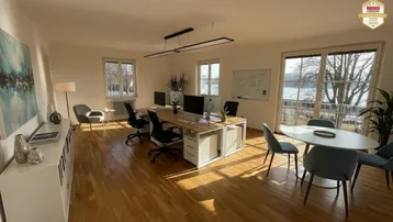 Ein helles, möbliertes Büro mit Holzboden, großen Fenstern und Blick auf das Wasser.