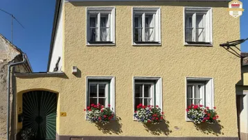 Gelbe Fassade eines traditionellen Gebäudes mit weißen Fensterläden und Blumenkästen unter blauem Himmel.