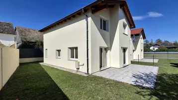Neubau-Doppelhaushälfte mit kleiner Rasenfläche und gepflasterter Terrasse unter blauem Himmel.
