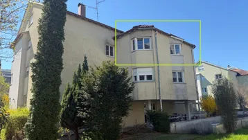 Mehrfamilienhaus mit beiger Fassade, Erker und gepflegtem Gartenbereich unter blauem Himmel.