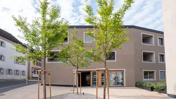 Modernes Gebäude mit beiger Fassade, vielen Fenstern und kleinen Bäumen davor, an einer Straße gelegen.