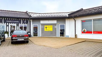 Außenansicht eines Gewerbegebäudes mit mehreren Ladenfronten und einem großzügigen Parkplatz davor.