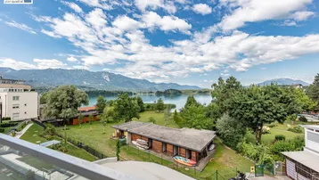 Panoramablick von der Terrasse auf den See, die umliegende grüne Landschaft und weitere Gebäude.