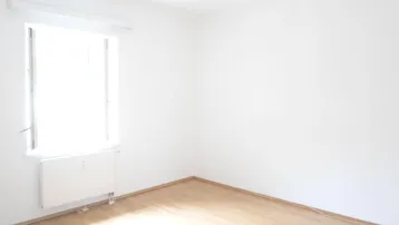 Heller, leerer Raum mit weißen Wänden, Holzfußboden und einem Fenster mit Heizkörper.
