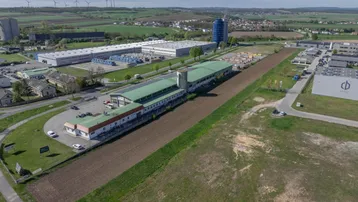 Luftaufnahme eines Gewerbegebiets mit mehreren Hallen, einem Ackerstreifen und Windrädern im Hintergrund.