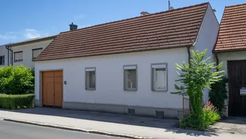 Vorderansicht eines kleinen, weiß verputzten Hauses mit braunem Ziegeldach und integrierter Garage.