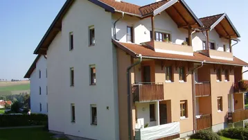 Mehrfamilienhaus mit mehreren Balkonen und Satteldach in ländlicher Umgebung, umgeben von Grünflächen.