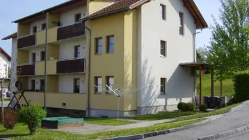 Mehrfamilienhaus mit mehreren Balkonen, einem kleinen Spielbereich und grüner Umgebung.