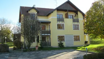 Gelbes Mehrfamilienhaus mit mehreren Balkonen, umgeben von Bäumen und einer Parkfläche.