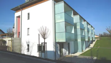Modernes Mehrfamilienhaus mit weißer Fassade, großen Fenstern und Glasbalkonen in grüner Umgebung.