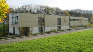Modernes Gebäude mit heller Fassade und großen Fenstern, umgeben von weitläufigen Grünflächen.