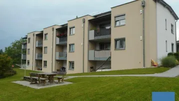 Modernes Mehrfamilienhaus mit beiger Fassade, Balkonen und einem gepflegten Gemeinschaftsgarten mit Sitzgelegenheit.
