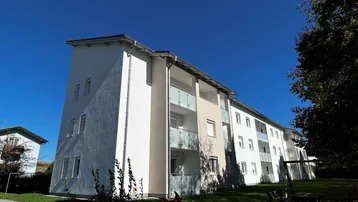 Modernes Mehrfamilienhaus mit weißer Fassade, Balkonen und gepflegter Grünanlage unter blauem Himmel.