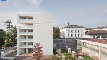 Modernes weißes Gebäude mit mehreren Balkonen und einer gepflegten Grünfläche im Vordergrund unter klarem Himmel.