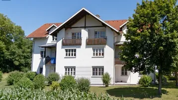 Weißes Mehrfamilienhaus mit Satteldach und Balkonen, umgeben von grüner Landschaft unter blauem Himmel.